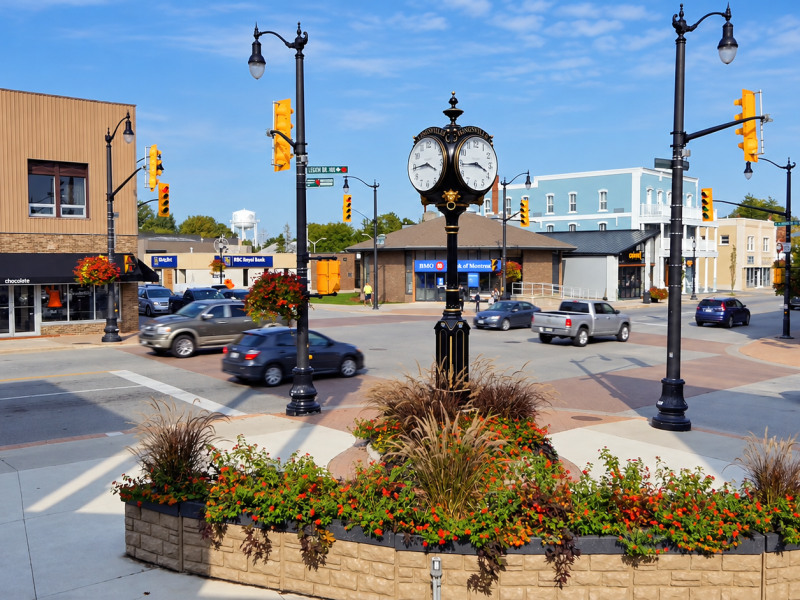Decorative Kingsville town clock above a flower-filled planter, with storefronts, street lamps, and traffic at the downtown intersection.